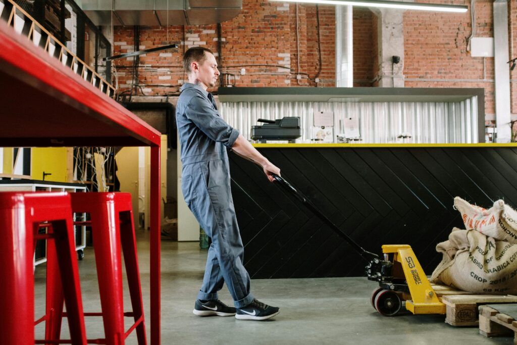 A man in overalls operates a pallet jack in a modern industrial loft with coffee sacks.