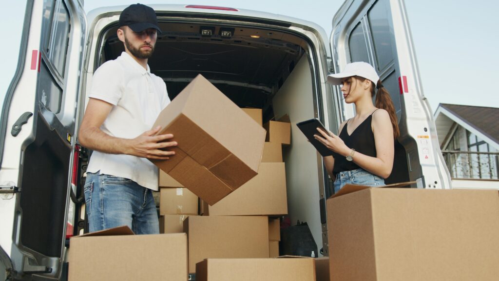 Two delivery workers handling cardboard boxes by a van, checking orders on tablet.